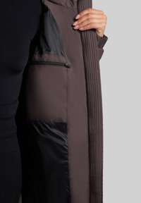 Notranjost rjave jakne s prešitjem prikazuje črn žep na zadrgo z gladko teksturo; tkanina se zdi lahka in izolirana.