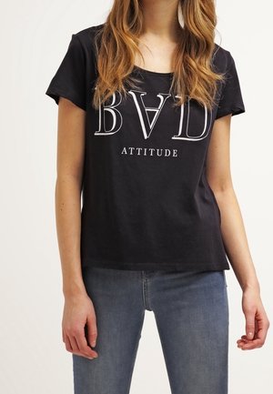 Femme portant un t-shirt noir avec l’inscription "BAD ATTITUDE" et un jean bleu, debout devant un fond clair uni.