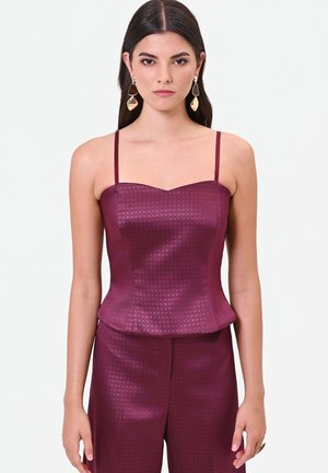 Donna con capelli lunghi e scuri indossa un top viola senza maniche texturizzato e pantaloni coordinati, accessoriati con grandi orecchini dorati e un anello.