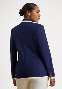 Moteris tamsia oda ir pynėmis surištu kuodu, dėvinti tamsiai mėlyną švarką su baltais apvadais ir auksiniais mygtukais, stovinti priešais šviesų vienspalvį foną.