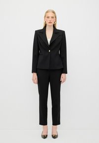 Zwarte op maat gemaakte kostuum met een enkele gouden knoop, gestructureerde schouders en slim-fit broeken. Glanzende zwarte schoenen met gouden accenten.