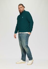 Teal sweatshirt met capuchon en klein logo, gecombineerd met blauwe spijkerbroeken en witte sneakers. Gladde textuur, relaxte pasvorm.