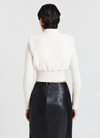 Gilet corto bianco in pelliccia sintetica abbinato a una gonna in pelle nera. Il gilet presenta maniche lunghe a coste e un colletto alto. Tessuto testurizzato visibile.