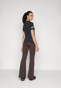 Svart kortermet skjorte med hvit "BOLD" tekst, kombinert med tettsittende brune flare-bukser, begge laget av glatt stoff. Modell vender bort.