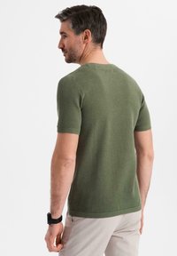 Suéter de punto verde oliva de manga corta y cuello redondo con una textura suave y un patrón de panal, combinado con pantalones claros y un reloj negro.
