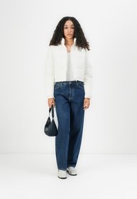 Witte cropped pufferjack met een rits aan de voorkant, gecombineerd met hoge blauwe denim jeans en lichtgekleurde schoenen. Houdt een kleine zwarte handtas vast.