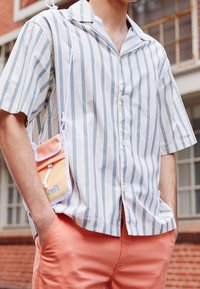 Camicia a maniche corte a righe bianca e blu, abbinata a pantaloni corti color corallo. Una piccola borsa a tracolla arancione è indossata in vita.