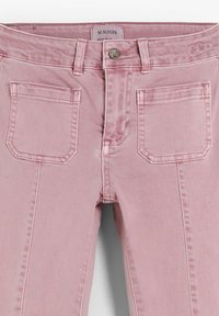 Jeans de mezclilla rosa claro con cintura alta, dos bolsillos traseros con esquinas redondeadas y cierre de botón de metal. Etiqueta de marca visible en el interior.