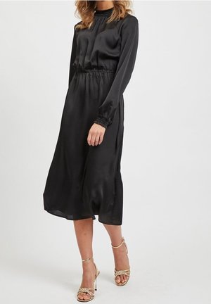 Kobieta ubrana w czarną sukienkę midi z długim rękawem, z marszczonym stanem i mankietami, w połączeniu ze złotymi sandałami na wysokim obcasie z paskami.