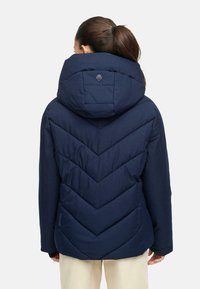 Temno moder puhovnik z oblikovanjem v obliki chevrona in velikim kapuco. Ima logotip na hrbtni strani in gladko teksturo.