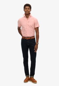 Camicia rosa a maniche corte con colletto e tasca, abbinata a jeans blu scuro e scarpe in pelle marrone. La cintura è marrone con una fibbia argentata.