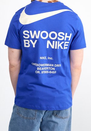 Niebieska bawełniana koszulka z dużym białym logo Nike na plecach, z napisem "SWOOSH BY NIKE" oraz adresem, nadrukowanym pogrubioną białą czcionką.