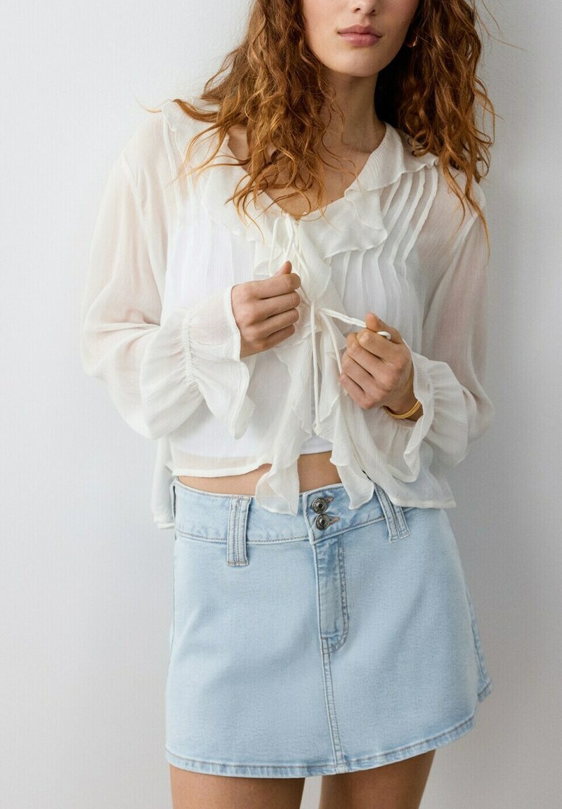 Femme portant un chemisier blanc transparent à volants, noué à l'avant, et une mini-jupe en denim bleu clair avec double boutons.