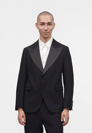 Man with shaved head wearing a black tuxedo jacket with satin lapels and a white dress shirt, standing against a plain white background.