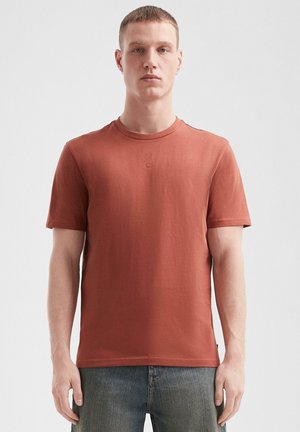 Jonge man met kort haar, gekleed in een roestkleurig T-shirt met korte mouwen en blauwe jeans, staande tegen een effen lichte achtergrond.