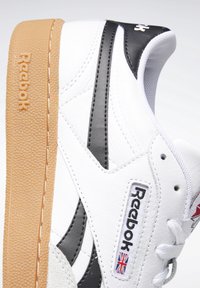 Bílé kožené tenisky s černými akcenty, texturovanou gumovou podrážkou, brandem Reebok a kulatými tkaničkami. Vyznačují se hladkým svrškem a prošívanými detaily.