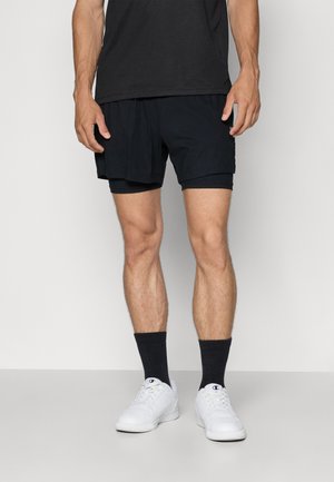 Shorts de sport noirs avec un design léger, présentant un aspect superposé et une texture lisse, associés à des baskets blanches et des chaussettes noires.