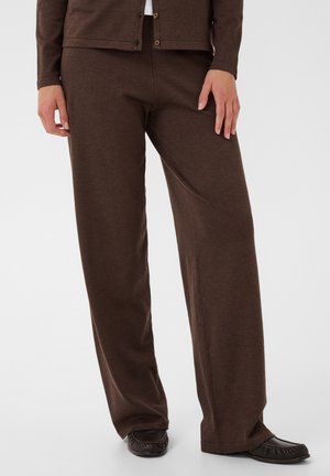 Personne portant un pantalon marron ample, un cardigan assorti boutonné et des chaussures marron, debout avec une main le long du corps.