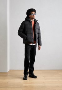 Czarna kurtka puchowa z kapturem, pomarańczowy sweatshirt pod spodem, czarne dżinsy i czarne buty. Model stoi na tle jasnoszarym.