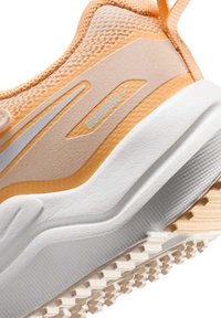 Zapatilla de correr con parte superior en tonos durazno y beige claro, superposiciones texturizadas, suela blanca con patrón de dibujo distintivo y detalles plateados.