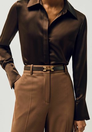 Cămașă din satin maro închis cu mâneci lungi băgată în pantaloni maro deschis cu talie înaltă, cu o curea neagră prevăzută cu o cataramă aurie.
