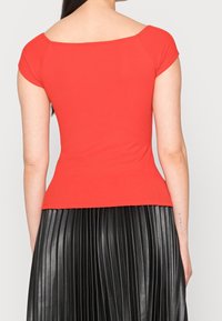 Top rosso attillato a coste con maniche corte, scollatura ampia e vita stile peplo, indossato con una gonna plissettata nera.