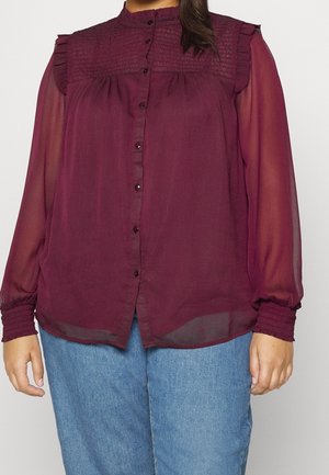 Mujer vestida con una blusa transparente burdeos con pechera y puños con volantes, cierre frontal con botones, combinada con vaqueros azul claro, sobre un fondo liso.