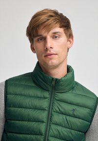 Grüne Steppweste mit hohem Kragen, versehen mit einer gesteppten Textur und einem zentralen Reißverschluss. Ein kleines Logo ist auf der linken Seite eingestickt.