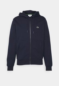 Sudadera con cremallera en color marino, con bolsillo tipo canguro, capucha con cordón y pequeño logotipo de cocodrilo verde en el lado izquierdo del pecho. Tela suave y texturizada.