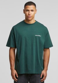 Tumši zaļš kokvilnas T-krekls ar īsām piedurknēm, apaļu dekoltē un priekšējā kreisā krūtī esošu logo ar uzrakstu "Cits karikatūra!" baltā krāsā.