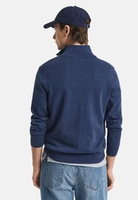 Marineblaues Strickpullover mit hohem Kragen, gerippten Bündchen und Saum, kombiniert mit hellblauen Jeans. Rückansicht, lässige Passform.
