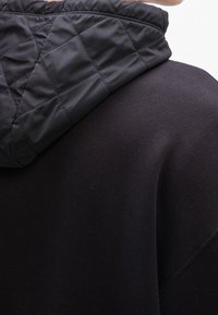 Gros plan d'une personne portant une veste noire à capuche avec un motif matelassé sur la capuche et un tissu lisse sur le corps.