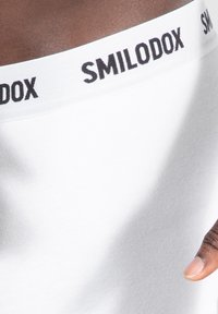 Top bianco senza maniche con una texture morbida e una larga fascia elastica nera con la scritta "SMILODOX" in lettere in grassetto.