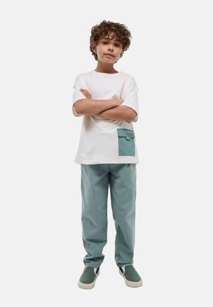 Garçon aux cheveux bouclés debout, bras croisés, portant un t-shirt blanc avec une poche bleue, un pantalon bleu et des chaussures vertes sans lacets sur fond blanc.