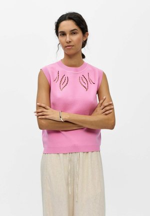 Frau mit dunklen Haaren, die ein pinkfarbenes ärmelloses Oberteil mit ausgeschnittenen Blattmustern und beige Kordelhosen trägt, steht mit verschränkten Armen.