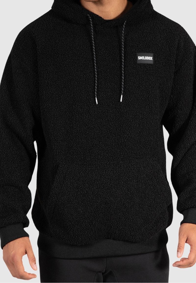 Svart fleecehoodie med en texturerad yta, justerbara dragsko, och en liten logotyp på bröstet. Ribbad muddar och nederkant.