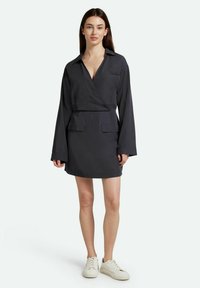 Dunkelgraues Wickelkleid mit langen Ärmeln, einem kragenlosen Ausschnitt, zwei aufgesetzten Taschen und einem geraden Saum. Kombiniert mit weißen Turnschuhen.