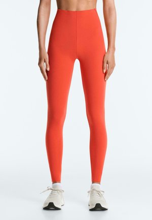 Colanți strâmți, cu talie înaltă, de culoare portocaliu aprins, purtați cu șosete albe până la gleznă și pantofi sport albi, văzuți de la talie până la picioare.