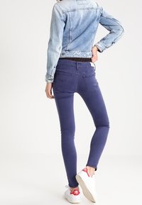 Hängselbyxor i mörklila denim, tillsammans med en ljusblå denimjacka. Vita sneakers med röda accenter kompletterar outfiten.