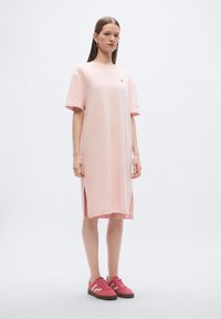 Φόρεμα από βαμβακερό ύφασμα σε ανοιχτό ροζ χρώμα με κοντά μανίκια, πλευρικές σχισμές και μικρό λογότυπο στο στήθος. Συνδυασμένο με ροζ αθλητικά παπούτσια με καφέ σόλες.