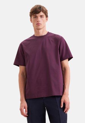 Giovane uomo con capelli castani corti che indossa una maglietta viola scuro tinta unita e pantaloni scuri, in piedi davanti a uno sfondo grigio neutro.