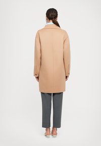 Donna con capelli scuri raccolti in una coda bassa, che indossa un cappotto color cammello, pantaloni grigi a vita alta e scarpe con tacco beige, in piedi di spalle su uno sfondo bianco.
