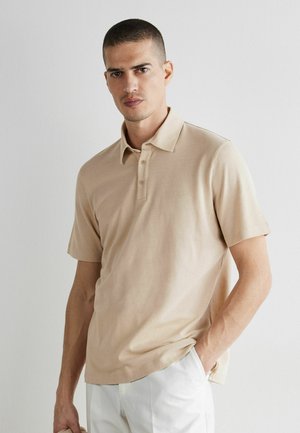 Mand med kort hår iført beige poloshirt med korte ærmer og hvide bukser, stående med den ene hånd i lommen mod en ensfarvet væg.