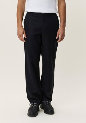 Man draagt zwarte broek met rechte pijpen, zwarte veterschoenen en een wit mouwloos topje, staand tegen een effen achtergrond.