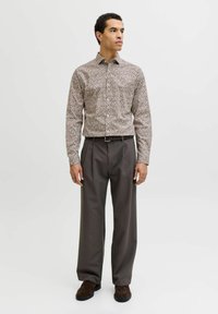 Homme debout portant une chemise beige à motifs floraux boutonnée, un pantalon plissé gris foncé, une ceinture noire et des chaussures marron foncé, regardant sur le côté.