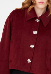 Burgundy cropped jacket with a soft texture, featuring a pointed collar and five silver-button closures along the front.