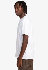 Camiseta de algodón blanca con mangas cortas, cuello redondo y un corte holgado, combinada con pantalones marrones. Diseño minimalista sin estampados.