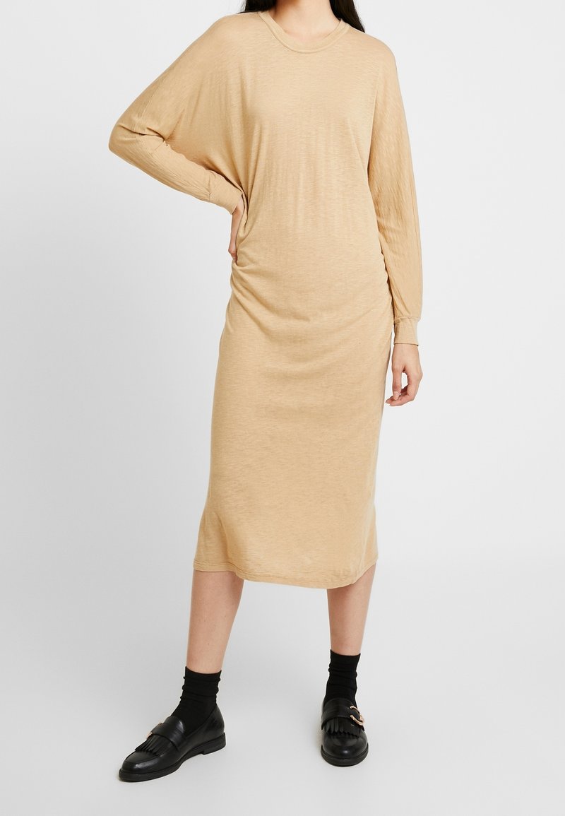 Beige midi-jurk met lange mouwen gemaakt van zachte, lichtgewicht stof met een losse pasvorm en een geplooid detail aan de zijkant, gecombineerd met zwarte loafers en sokken.