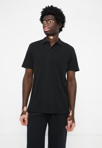 Fekete pólóing, rövid ujjú, pamutból készült, klasszikus gallérral és diszkrét, dombornyomott logóval a mellkason.