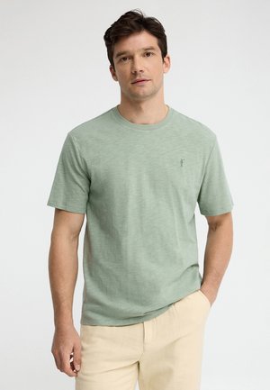 Uomo che indossa una maglietta verde chiaro a maniche corte e pantaloni beige, in piedi con una mano in tasca contro uno sfondo semplice.